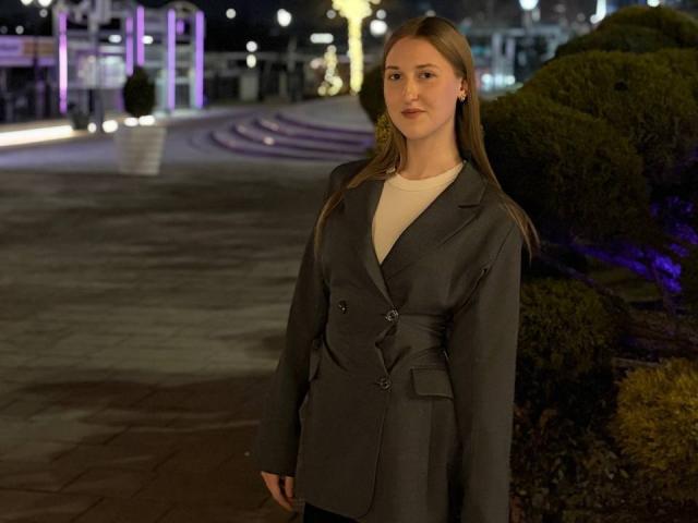 NaSaxe está em um ambiente externo noturno, vestindo um blazer cinza de corte duplo sobre uma camisa branca, com cabelos castanhos longos e uma expressão neutra. Seus seios são pequenos..