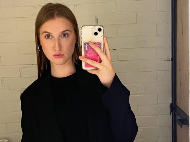 NaSaxe mit glattem, braunem Haar, trägt ein schwarzes Oberteil und eine Jacke, hält ein weißes Telefon mit einer rosa Hülle, blickt in einen Spiegel, trägt kleine Ohrringe im Stecker-Design, weiße Ziegelwand..