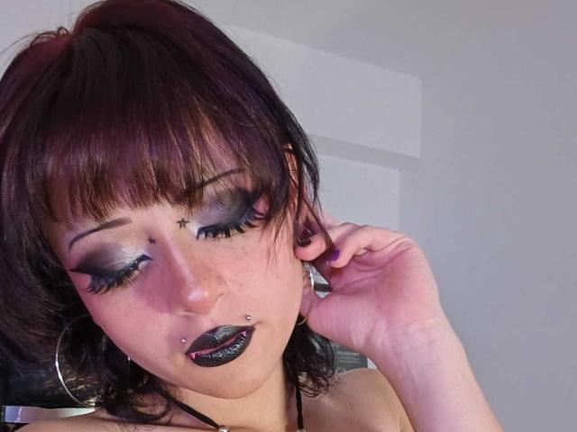 ViolettClay has dark purple hair, heavy black eyeliner, purple eyeshadow, black lipstick, a lip piercing, and a nose piercing. She's wearing hoop earrings and touching her hair.