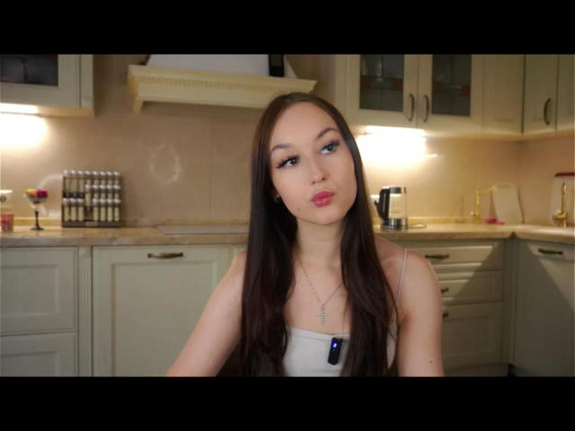 Anika Trentz, con cabello castaño largo, vestida con una camiseta blanca, frunciendo sus labios rosados, con un pequeño collar de cruz, en una cocina con luz tenue, mirando directamente a la cámara..