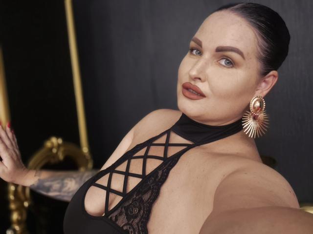 GeorgiaLerox, com cabelo escuro, batom vermelho e brincos de ouro, exibindo seus seios grandes em uma blusa preta com amarração, tatuagem no braço, segurando o celular para tirar uma selfie..