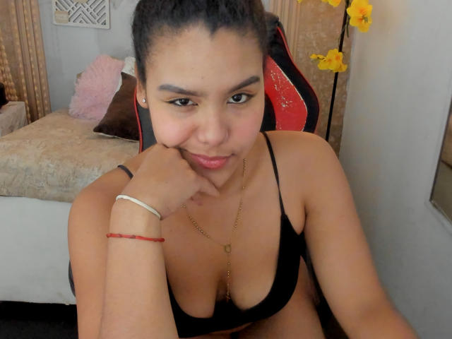 AriaVega, con i capelli scuri raccolti in una coda di cavallo, indossa un reggiseno nero, una collana dorata e un braccialetto rosso, sorride, appoggiando il mento sulla mano, mostrando piccoli seni, seduta su una sedia rossa e nera..