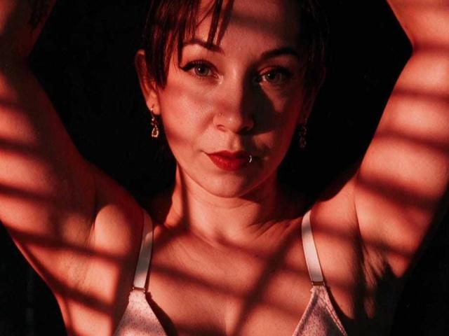 Sara Smith, con i capelli castano corti, le braccia alzate, indossa un reggiseno rosa, ha le labbra rosse, un piercing al naso, orecchini ed è illuminata da persiane. Il suo seno è visibile, ha la pelle chiara e un'espressione seria..