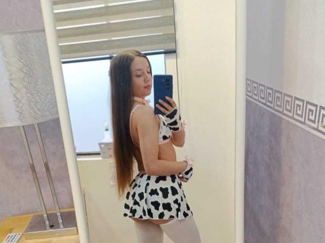 VioletaNova, avec de longs cheveux bruns, portant un soutien-gorge et une jupe à motifs de vaches, des bas blancs montants jusqu'aux cuisses, et des gants à motifs de vaches, prend un selfie devant un miroir, montrant ses fesses et son dos..