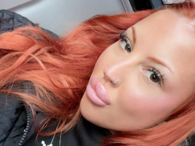 Roxyfire, con cabello rojo, un piercing en la nariz y lápiz labial rosa, frunciendo sus labios brillantes. Lleva una chaqueta negra acolchada y tiene pestañas largas y una ligera sonrisa..