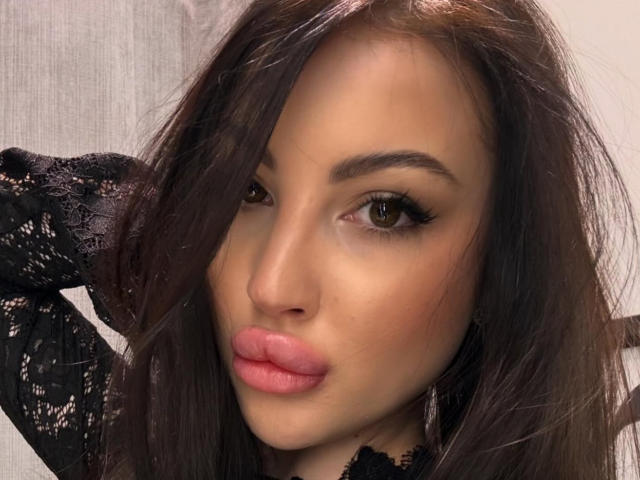 Le visage de Roxyfire, avec ses yeux marron, son rouge à lèvres rose et ses longs cheveux bruns, portant un haut en dentelle noire, regardant légèrement sur le côté avec une main dans les cheveux..