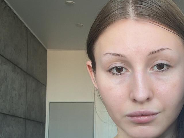 LailaBonanno hat ein blasses, mit Sommersprossen übersätes Gesicht mit braunen Augen, glattem, braunem Haar und rosafarbenen Lippen. Sie blickt leicht zur Seite und hat einen neutralen Gesichtsausdruck..