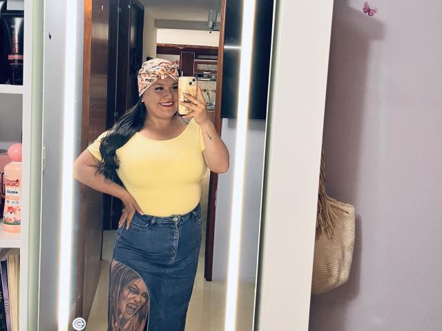 Colette Bessette, avec de longs cheveux noirs, un haut jaune, un jean taille haute, un bandana fleuri, un tatouage sur la cuisse droite, souriante, tenant un téléphone dans une photo de profil prise dans un miroir, une main sur la hanche..