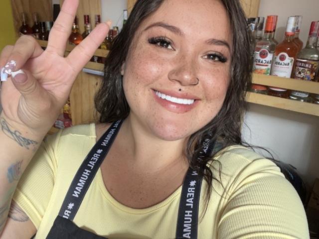 Colette Bessette, con i capelli castani, lentiggini e un piercing al naso, sorride mostrando i denti bianchi, indossa una camicia gialla, un grembiule nero e fa un segno di pace, mostrando un tatuaggio..