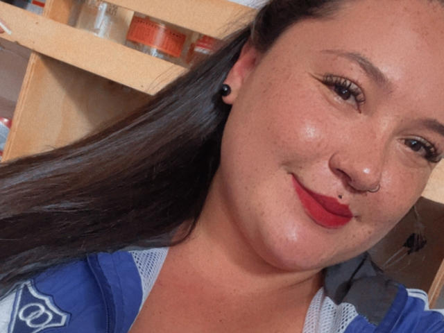 ColetteBessette tem seios médios, um piercing no nariz, brincos pretos, batom vermelho e cabelos castanhos longos, sorrindo com maquiagem discreta, vestindo uma jaqueta esportiva azul e branca..