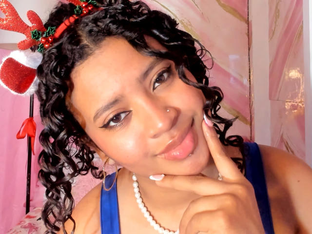 JessyZoee mit lockigem, schwarzem Haar, die ein rotes und weißes Stirnband, ein blaues Kleid, eine weiße Perlenkette und goldene Ohrringe trägt, steckt ihre Zunge heraus und berührt ihre Wange..