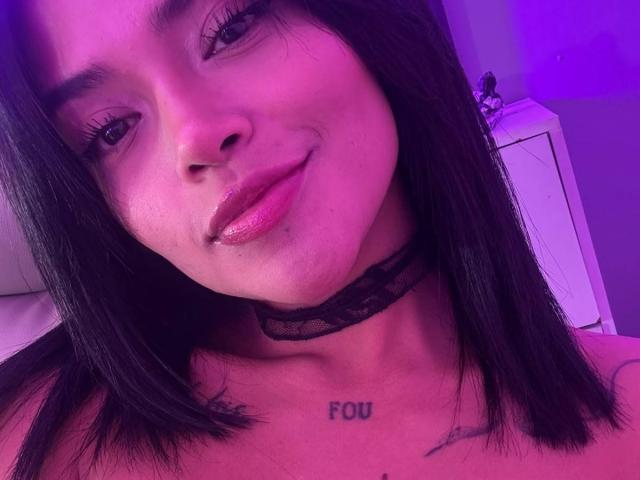 MajoJones con un collar de encaje negro, labios rosados brillantes, cabello negro lacio, tatuaje "FOU" en el pecho, una sonrisa sutil, iluminación púrpura, clavículas visibles y un tatuaje pequeño en el lado derecho..