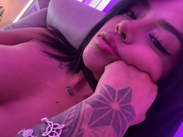 MajoJones con labios rosados brillantes, cabello negro, guiñando un ojo, con el mentón apoyado en un brazo tatuado, mostrando un tatuaje de estrella y una pulsera, bajo una luz púrpura, sin ropa, mostrando un pequeño tatuaje debajo del mentón..