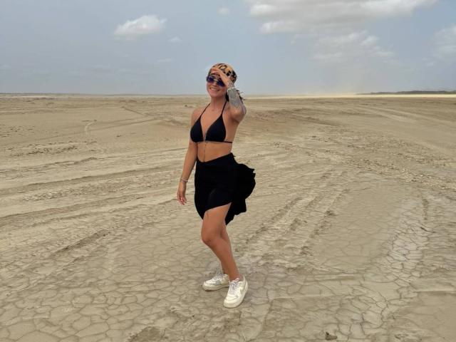 MajoJones, con piel bronceada, top de bikini negro, falda negra, zapatillas blancas y gafas de sol, de pie sobre terreno desértico agrietado, sonriendo, con los brazos relajados, un tatuaje en el brazo derecho, cielo nublado..