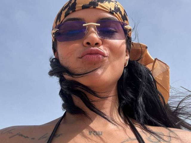 MajoJones, con cabello negro, usando un pañuelo de camuflaje, gafas de sol moradas con una barra dorada, labios fruncidos, tatuajes en el pecho y un accesorio de color bronceado, similar al cuero, en la cabeza..
