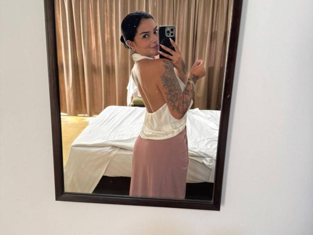MajoJones, con tatuajes, vestida con una camiseta blanca sin mangas y una falda rosa, tomando una foto de sí misma en un espejo, mostrando su espalda y trasero, en una habitación con una cama y una cortina..