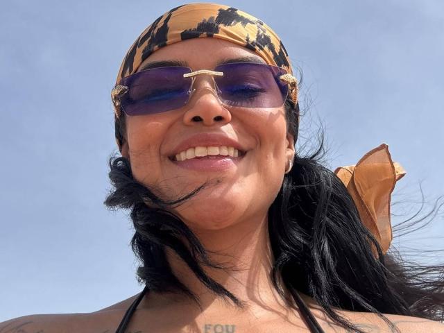 MajoJones, con un pañuelo de cabeza con estampado en tonos marrones y negros, gafas de sol moradas y cabello negro, sonriendo y mostrando tatuajes visibles en el pecho, lleva un lazo amarillo en el lado derecho..