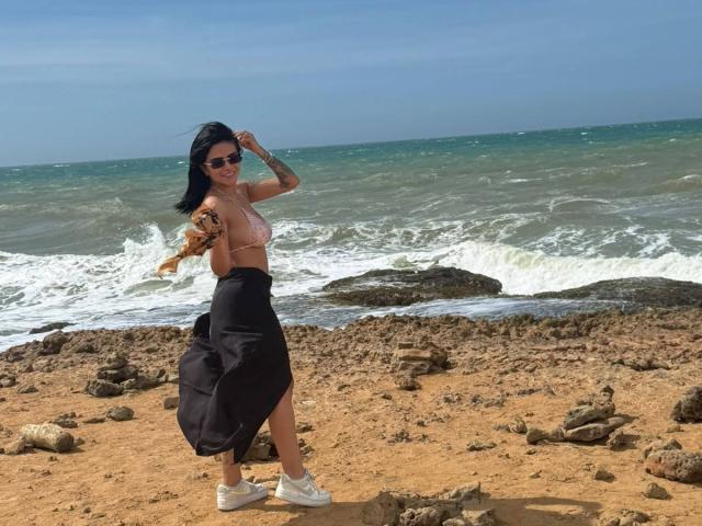 MajoJones, con cabello negro, gafas de sol y un tono de piel bronceado, de pie en una playa rocosa, vestida con un sujetador rosa y una falda negra, sosteniendo un perro, con zapatillas blancas..