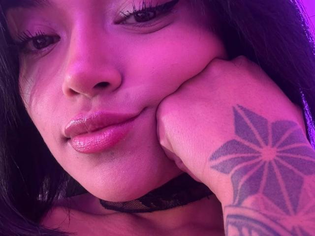 El rostro de MajoJones, con labios rosados y brillantes, un collar negro y una mano tatuada apoyada en su mejilla. La iluminación púrpura resalta su piel lisa y su cabello oscuro y ondulado..