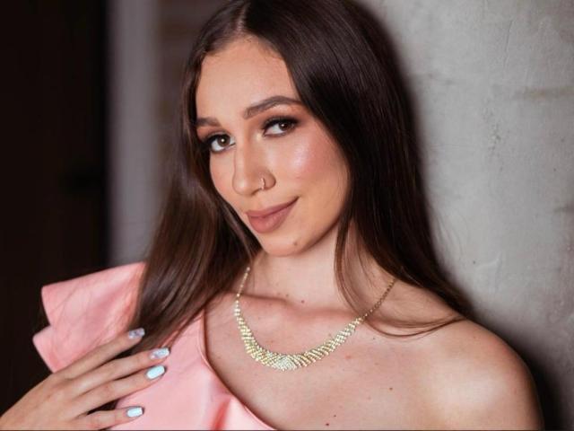 Maittee, met lang bruin haar, een neusring en een subtiele glimlach, draagt een gouden ketting en een roze top, waarbij ze haar borsten laat zien en lichte blauwe nagels heeft..