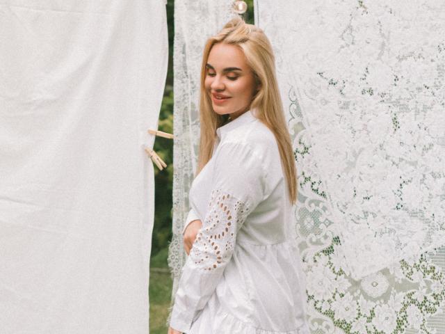 Gabriella Martins, con cabello rubio, vestido de encaje blanco y mangas bordadas, de pie cerca de cortinas de encaje blanco, sonriendo suavemente, mirando hacia abajo. Se ven los senos a través del vestido. Luz suave. Entorno al aire libre..