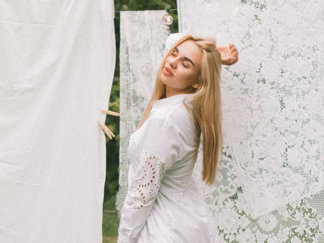 Gabriella Martins mit blonden Haaren, die Augen geschlossen hat, trägt ein weißes Spitzenkleid und steht vor einer mit Spitze bedeckten Wand. Ihre Hände sind erhoben, und sie trägt Holz-Wäscheklammern auf dem Kleid..
