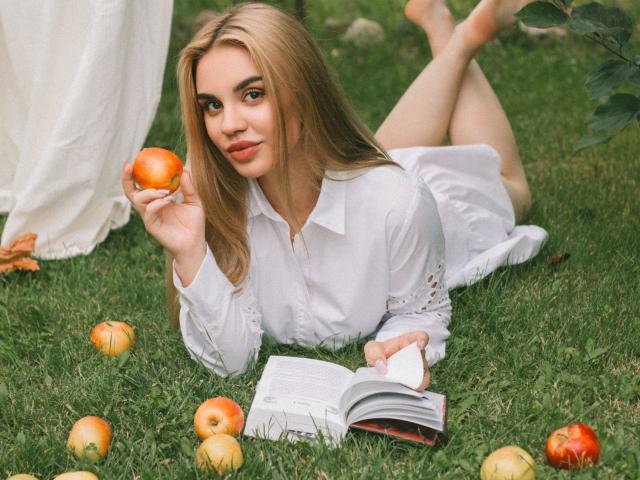 Gabriella Martins, com cabelo loiro, deitada na grama, vestindo um vestido branco, segurando uma maçã, com as pernas levantadas, lendo um livro, com maçãs ao redor, sorrindo, segurando uma maçã na mão, pés descalços, gramado verde..