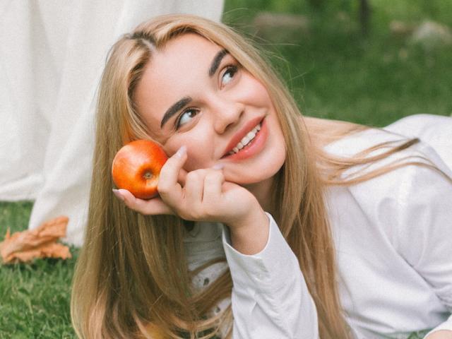 Gabriella Martins, com cabelos loiros longos, camisa branca, segurando uma maçã vermelha perto da bochecha, sorrindo para cima, grama verde, tecido branco ao fundo, folhas de outono, luz natural suave..