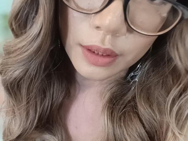 El rostro de TransKathie, con cabello castaño ondulado, labios rosados y gafas de montura gruesa, muestra una ligera sonrisa. Su nariz es pequeña y tiene un tono de piel claro..