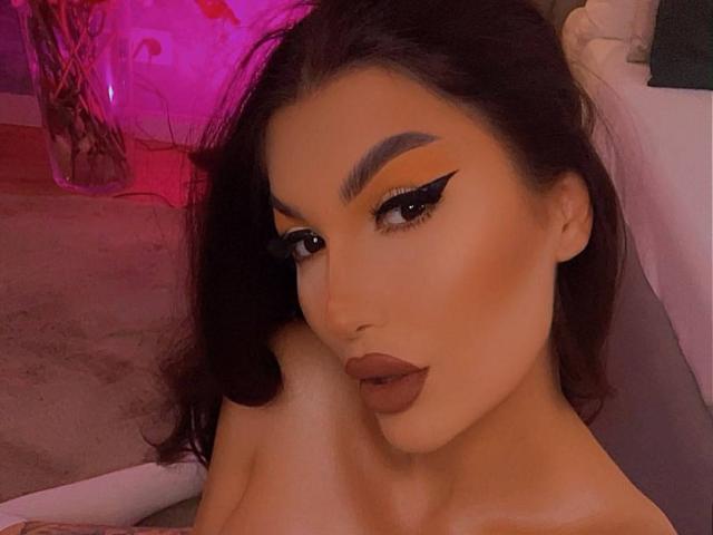 C'est JessieBabe, avec des sourcils épais et foncés, un trait d'eye-liner noir net, un fard à paupières orange et un rouge à lèvres marron. Elle a de longs cheveux bruns ondulés et une expression neutre. Ses seins sont légèrement visibles..