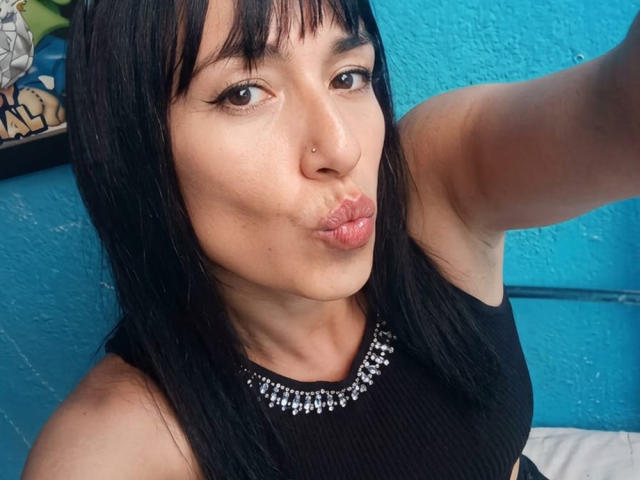 SyntiaLm mit schwarzen Haaren, Nasenpiercing und schwarzem Tanktop mit silberner Halskette, die ihre Lippen zu einem Schmollmund formt, auf einem Selfie. Ihre Brüste sind nicht sichtbar. Blaue Wand im Hintergrund..