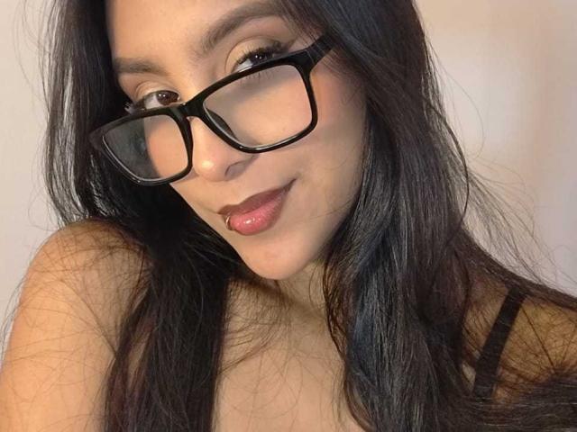 EmmaEstreilla, avec ses longs cheveux noirs, ses lunettes noires et un léger sourire, laissant entrevoir un piercing sur la lèvre, porte un haut noir à bretelles et affiche un regard confiant..