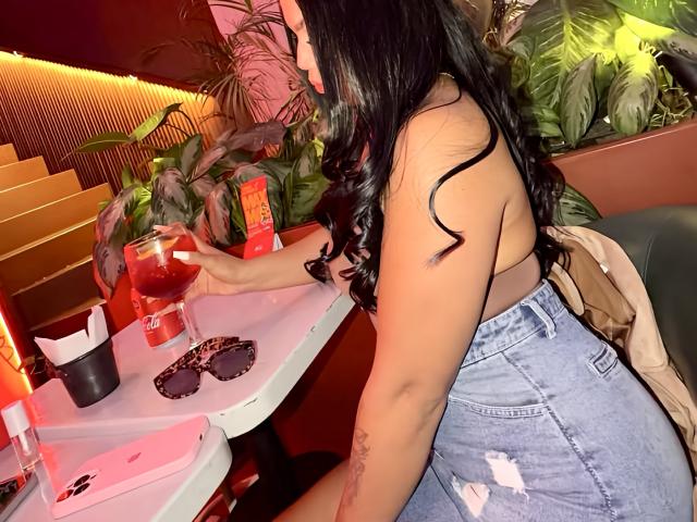 MiaDenver with long black hair, topless, showing her tits, wears a ripped blue denim skirt, sits at a table with a red drink, sunglasses, and a bag.