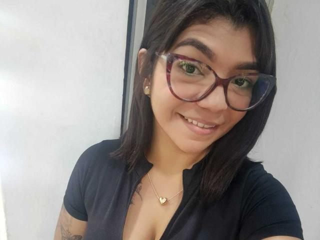 ValeryPierce with dark brown hair, green eyes, and red glasses, smiling, wearing a black top, heart necklace, and small earrings, showing tattoos on her arms.