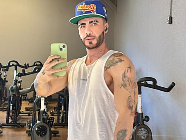 LancelotBenwick, met tatoeages, draagt een wit tanktop en een blauwe pet, en maakt een selfie in een sportschool bij een fietsapparaat, terwijl hij een groene telefoon vasthoudt en naar de camera kijkt..