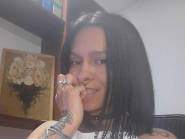 AmelieBlake with black hair, nose ring, smiling, white tank top, choker, ring on finger, tattoo on hand, holding finger to mouth, shelf with framed art in background.
