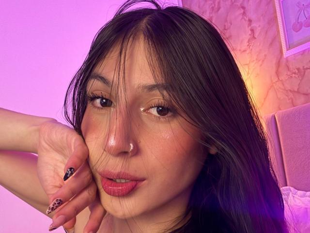 Sophie Beaumont, met donker haar, roze lippen, een neusring, bruine ogen en verzorgde nagels met zwarte en gouden patronen, die haar wang aanraakt in een selfie met paars licht..