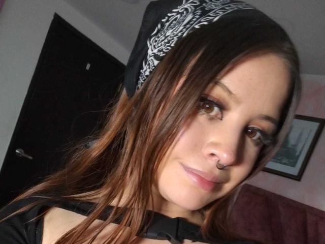Melani Becker, com cabelo castanho, lenço de cabeça preto e branco, piercing no nariz, sorriso discreto e maquiagem leve, olhando para a câmera, em um quarto com pouca luz..