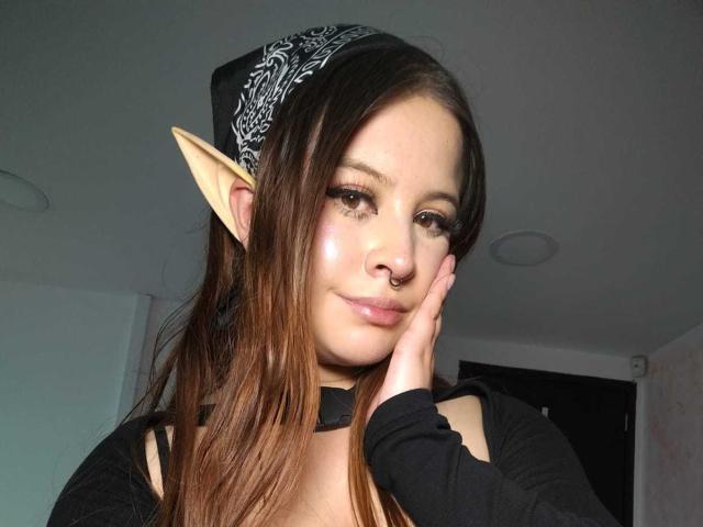 MelaniBecker with elf ears, brown hair, black bandana, nose ring, and shiny face, touching cheek, wearing black top, in selfie.
