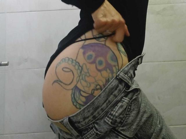 Il sedere di ZarayFeet, con un tatuaggio colorato di un polpo, un perizoma nero e dei jeans grigi tirati giù, mostra una mano che afferra l'elastico..