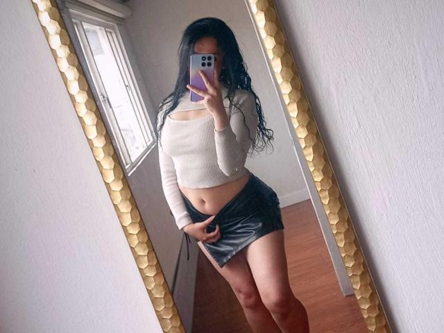 SharonKm, avec ses longs cheveux noirs, son crop top blanc et sa jupe en cuir noir, montrant sa vulve sous la jupe, prend un selfie devant un miroir, tout en tenant un téléphone violet..
