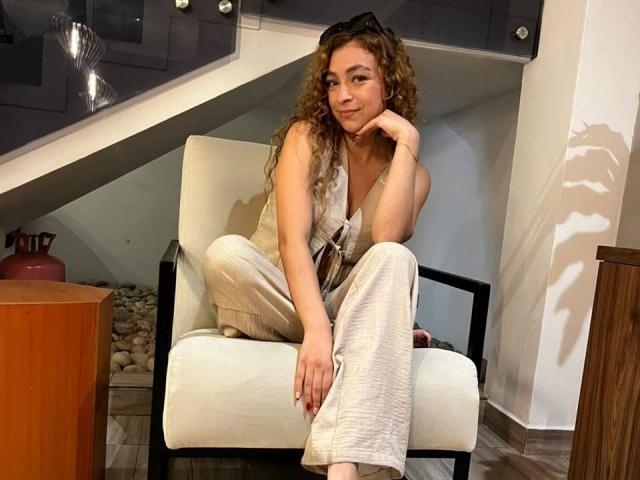 AriannaBlake with curly brown hair, sitting in a white chair, wearing a sleeveless beige top and white pants, hand on chin, sunglasses on head, smiling softly.