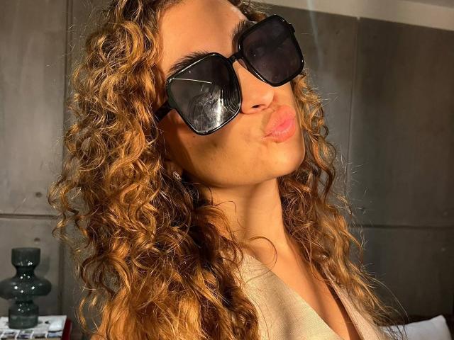 AriannaBlake with curly brown hair, pouty lips, and black oversized sunglasses, wearing a beige top, looking slightly to the side. Her nose is visible, and she's pouting.