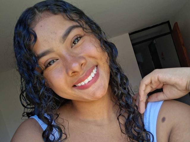 Lucia Toloza tiene el pelo negro y rizado, una sonrisa alegre que muestra los dientes blancos, y lleva una camiseta azul sin mangas, con una pequeña peca oscura en el hombro derecho..
