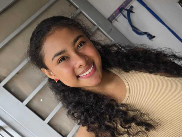 Lucia Toloza a les cheveux noirs bouclés, les yeux marron et un sourire qui révèle des dents blanches. Elle porte un débardeur beige texturé et des boucles d'oreilles rouges, et elle a l'air heureuse..