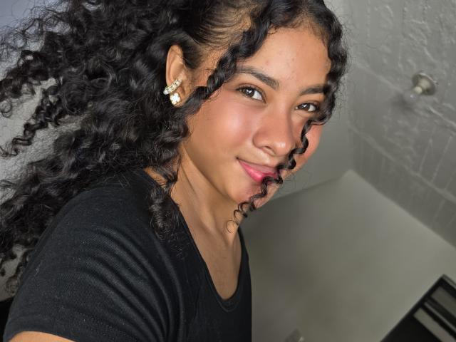 LuciaToloza tiene el pelo negro y rizado, piel de tono marrón medio y lleva una camisa negra. Está sonriendo, con lápiz labial rosa, mostrando su pequeña nariz y su cara redonda y linda..