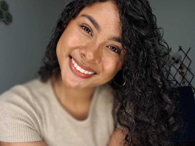 Lucia Toloza: Een vrouw met krullend zwart haar, bruine ogen en een glimlach waarbij witte tanden zichtbaar zijn, draagt een beige shirt en heeft een schattig gezicht en oorringen..