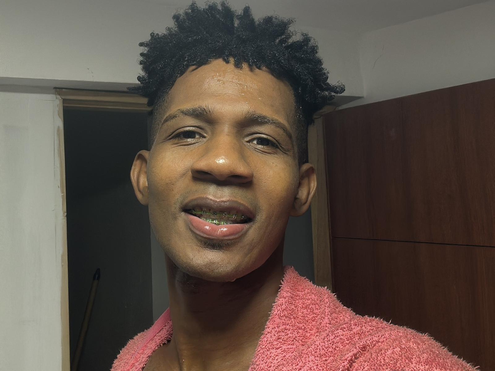 DanteFerro, com cabelo preto encaracolado e curto, pele escura e uma toalha rosa, sorrindo com um dente de ouro e um pequeno bigode, em um quarto com pouca luz..