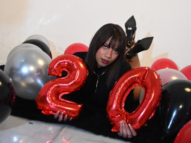 ZaraBlaze with dark brown hair, black outfit, holding shiny red "20" balloons, surrounded by black, red, and silver balloons, wearing black rabbit ears, smiling at camera.