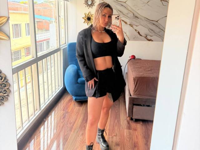 Elena Saenz, con piel morena y cabello rubio, vestida con un top corto negro, una falda corta, una chaqueta abierta y botas negras, está tomando una selfie frente al espejo, mostrando sus pechos y glúteos..